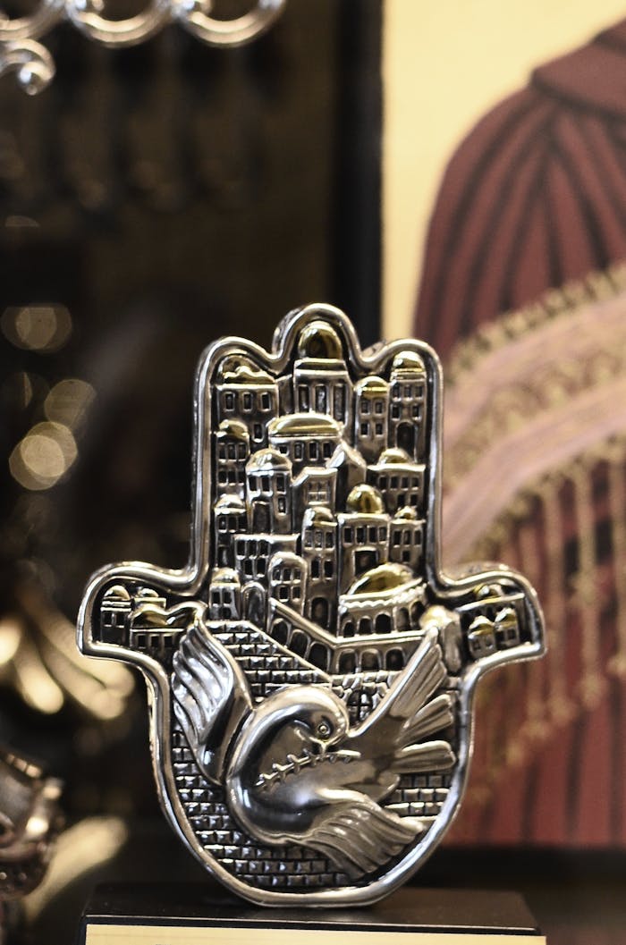 A decorative silver Hamsa featuring a bird and cityscape design, symbolizing protection and tradition.